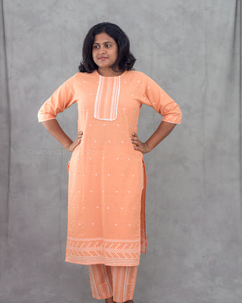 Woman wearing a peach-colored traditional outfit with white patterns against a gray background